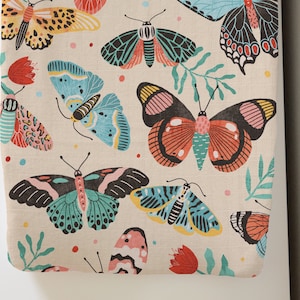 May include: A beige fabric with a repeating pattern of colorful butterflies. The butterflies are in various shades of blue, pink, red, yellow, and green. The fabric has a subtle texture.