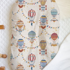 May include: A baby changing pad cover with a repeating pattern of colorful hot air balloons. The balloons are red, blue, yellow, and orange, with decorative flags and details. The background is a cream color with small speckles.