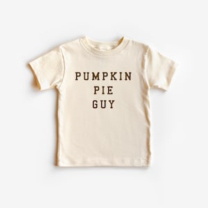 May include: Cream-colored t-shirt with the text "PUMPKIN PIE GUY" in dark brown block letters. The shirt is a short-sleeved crew neck style, likely for a child. The text is centered on the front of the shirt.
