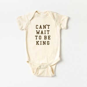 May include: Cream-colored baby bodysuit with the text "CAN'T WAIT TO BE KING" in dark brown block letters. The onesie has short sleeves and snaps at the bottom. A simple and sweet design for a baby.