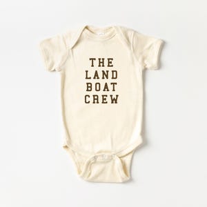 May include: Cream-colored baby bodysuit with the text "THE LAND BOAT CREW" in dark brown block letters. The one-piece garment has short sleeves and snaps at the bottom. The background is plain white.