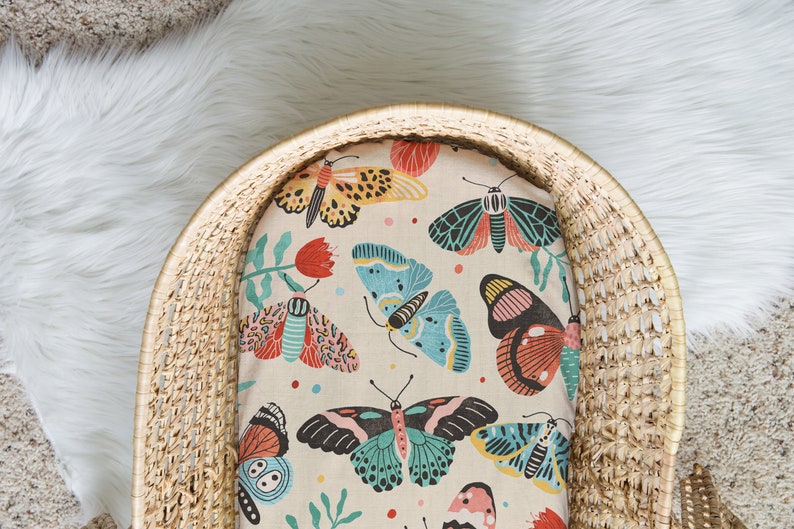 May include: A woven wicker basket with a cream-colored fitted sheet featuring a colorful butterfly and floral pattern. The sheet has a variety of butterflies in shades of blue, pink, red, and yellow.