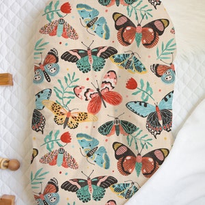 May include: A beige oval-shaped baby lounger with a colorful butterfly print. The print features butterflies in shades of pink, red, blue, green, and yellow on a beige background.