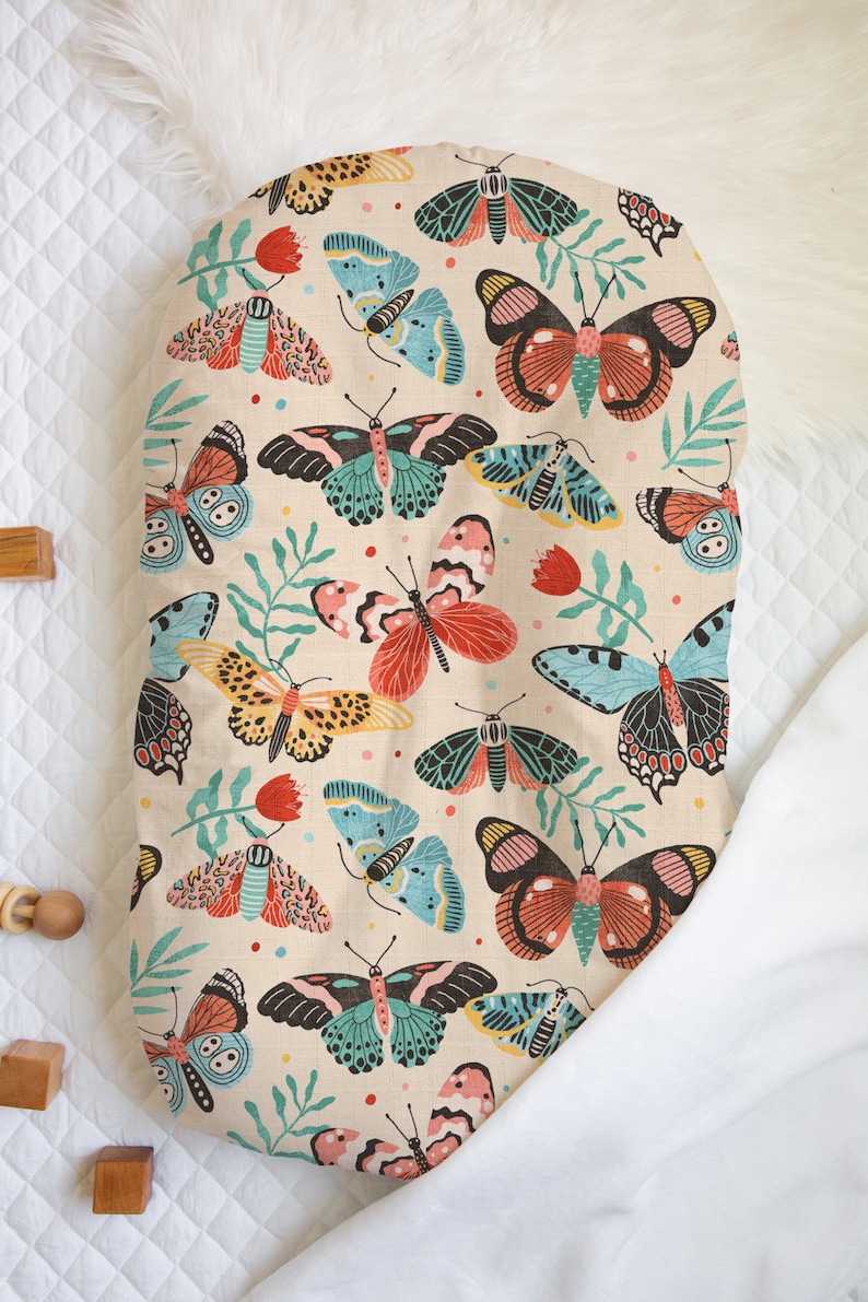 May include: A beige oval-shaped baby lounger with a colorful butterfly print. The print features butterflies in shades of pink, red, blue, green, and yellow on a beige background.