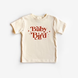 May include: A cream colored t-shirt with short sleeves and the text "Baby Bird" in orange lettering with small star accents.