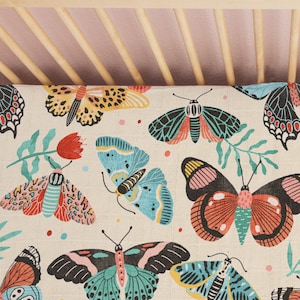 May include: A white crib sheet with a colorful butterfly pattern. The butterflies are in shades of blue, green, pink, red, and yellow. The sheet has a light beige background.