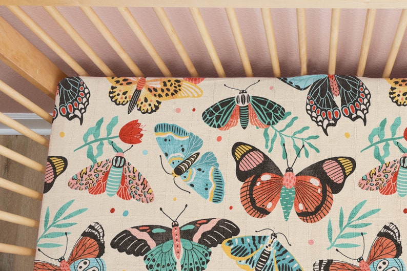May include: A white crib sheet with a colorful butterfly pattern. The butterflies are in shades of blue, green, pink, red, and yellow. The sheet has a light beige background.