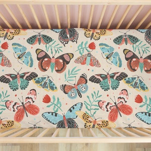 May include: A cream-colored fitted crib sheet with a colorful butterfly pattern. The butterflies are in shades of blue, green, red, and yellow. The sheet is made of a soft, breathable fabric.