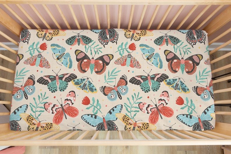 May include: A cream-colored fitted crib sheet with a colorful butterfly pattern. The butterflies are in shades of blue, green, red, and yellow. The sheet is made of a soft, breathable fabric.