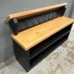 Puede incluir: Un banco de entrada de madera con asiento de madera natural clara y base negra con dos estantes abiertos. Un panel trasero incluye un estante superior de madera a juego y una fila de ocho ganchos de metal negro para colgar objetos.