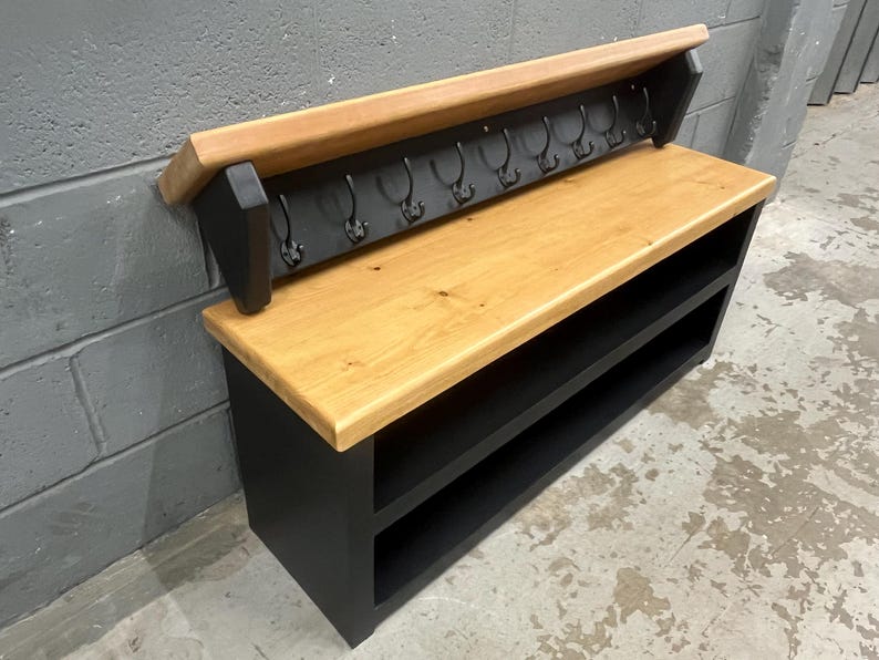 Puede incluir: Un banco de entrada de madera con asiento de madera natural clara y base negra con dos estantes abiertos. Un panel trasero incluye un estante superior de madera a juego y una fila de ocho ganchos de metal negro para colgar objetos.