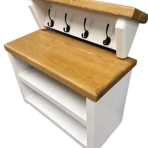 Puede incluir: Un banco de entrada de madera natural y blanca que cuenta con un estante superior con cuatro ganchos de metal negro. La unidad incluye un asiento de madera plano y dos estantes de almacenamiento abiertos debajo, con marco blanco y superficie te&ntilde;ida.