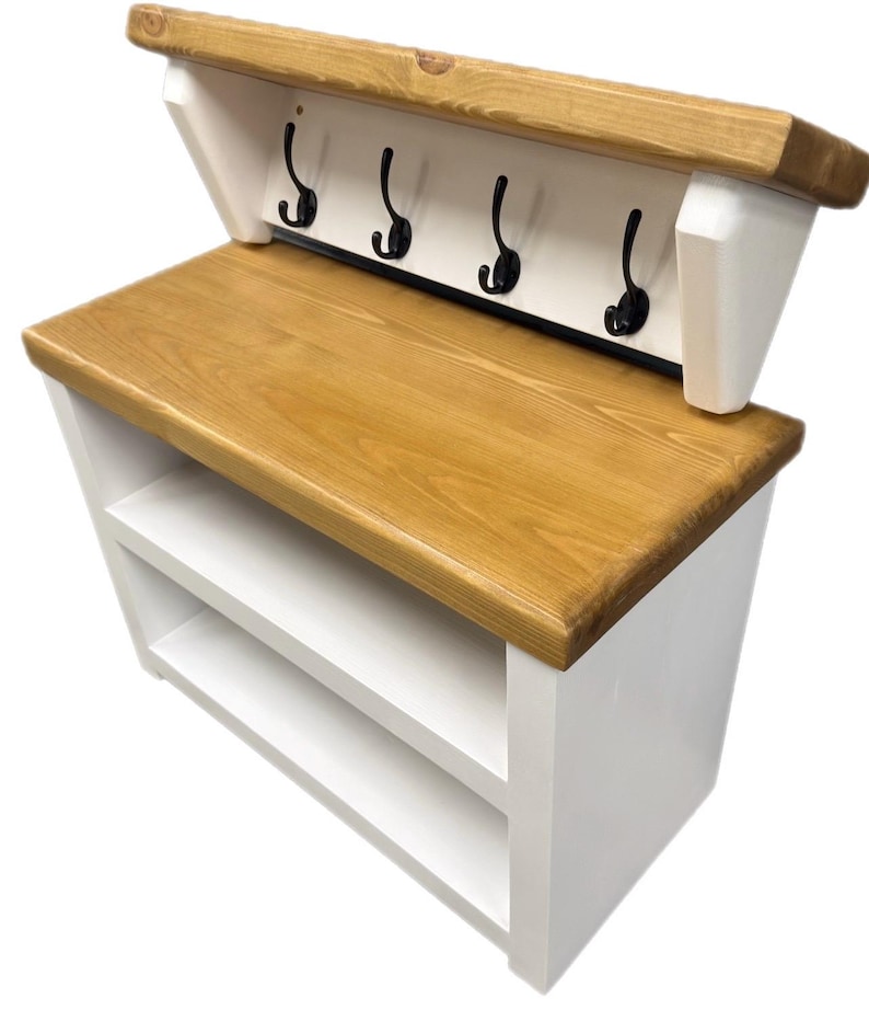 Puede incluir: Un banco de entrada de madera natural y blanca que cuenta con un estante superior con cuatro ganchos de metal negro. La unidad incluye un asiento de madera plano y dos estantes de almacenamiento abiertos debajo, con marco blanco y superficie te&ntilde;ida.