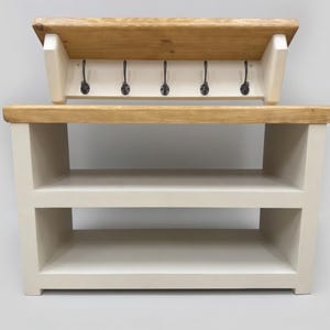 Puede incluir: Un banco de entrada de madera con una superficie de madera natural y una base de color crema con dos estantes abiertos. En la parte superior, un perchero de pared a juego cuenta con cinco ganchos de metal negro y una repisa de madera.