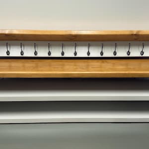 Puede incluir: Un banco de entrada de madera con asiento y estante superior en tono madera natural, contrastado por una estructura blanca. La unidad incluye dos estantes de almacenamiento abiertos en la base y un panel trasero con trece ganchos met&aacute;licos negros.