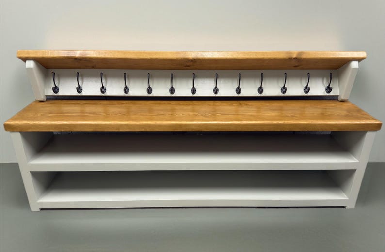 Puede incluir: Un banco de entrada de madera con asiento y estante superior en tono madera natural, contrastado por una estructura blanca. La unidad incluye dos estantes de almacenamiento abiertos en la base y un panel trasero con trece ganchos met&aacute;licos negros.