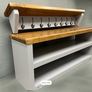 Puede incluir: Un mueble de recibidor con base gris claro, dos estantes abiertos y una secci&oacute;n superior a juego con tapa de madera natural. La parte superior incluye nueve ganchos de metal negro para colgar objetos, montados sobre un panel trasero gris claro.