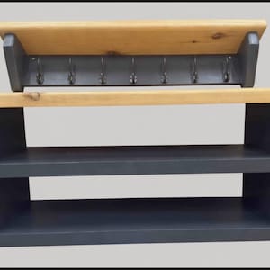 Puede incluir: Un zapatero de madera gris oscuro con dos estantes abiertos y una superficie superior de madera clara natural. Sobre el mueble hay un perchero de pared a juego en gris oscuro con un estante de madera clara y siete ganchos met&aacute;licos.