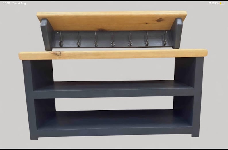 Puede incluir: Un zapatero de madera gris oscuro con dos estantes abiertos y una superficie superior de madera clara natural. Sobre el mueble hay un perchero de pared a juego en gris oscuro con un estante de madera clara y siete ganchos met&aacute;licos.