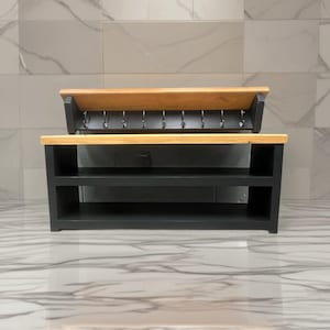 Puede incluir: Un organizador de entrada de madera negra con un estante superior de madera natural y un zapatero inferior con dos niveles abiertos. Sobre el zapatero, un perchero de pared incluye una parte superior de madera natural y ocho ganchos.