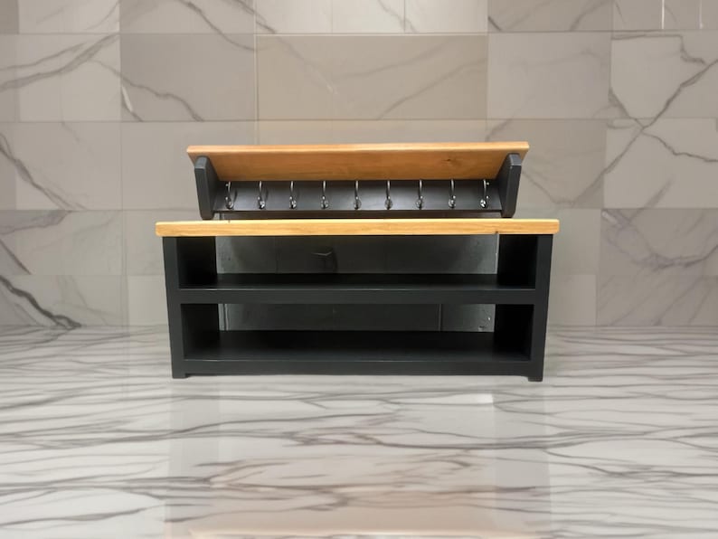Puede incluir: Un organizador de entrada de madera negra con un estante superior de madera natural y un zapatero inferior con dos niveles abiertos. Sobre el zapatero, un perchero de pared incluye una parte superior de madera natural y ocho ganchos.