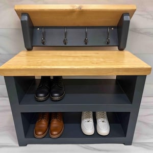 Puede incluir: Un banco recibidor con una superficie de madera natural y un marco gris oscuro. La unidad incluye un panel trasero con cuatro ganchos de metal negro y dos estantes inferiores para guardar zapatos. La estructura es rectangular y robusta.