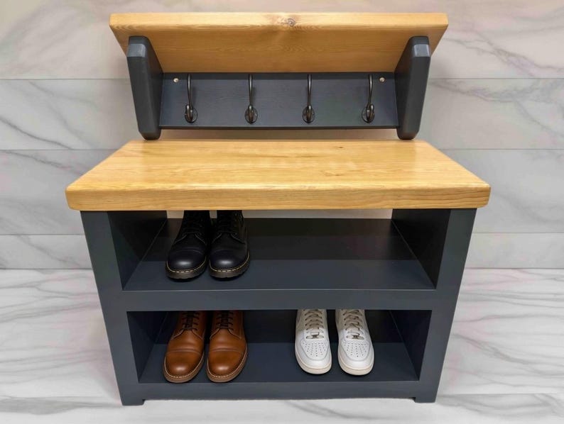 Puede incluir: Un banco recibidor con una superficie de madera natural y un marco gris oscuro. La unidad incluye un panel trasero con cuatro ganchos de metal negro y dos estantes inferiores para guardar zapatos. La estructura es rectangular y robusta.