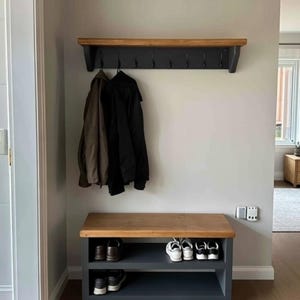 Puede incluir: Un perchero de pared con estante superior de madera natural y base gris oscuro con ocho ganchos de metal negro. Debajo, un banco zapatero a juego con superficie de madera natural y dos estantes abiertos de color gris oscuro para zapatos.