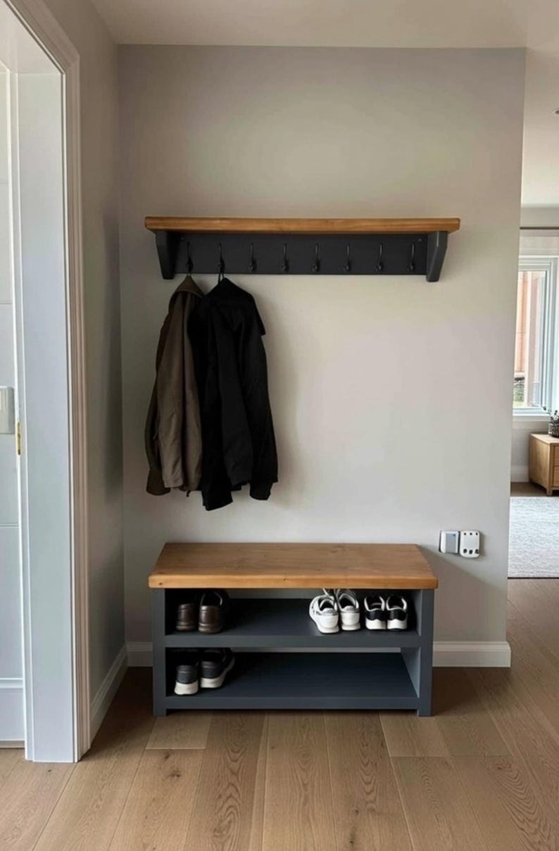 Puede incluir: Un perchero de pared con estante superior de madera natural y base gris oscuro con ocho ganchos de metal negro. Debajo, un banco zapatero a juego con superficie de madera natural y dos estantes abiertos de color gris oscuro para zapatos.