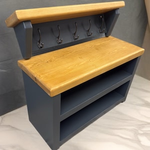 Puede incluir: Un organizador de entrada montado en la pared con un estante superior de madera natural y una base gris oscuro. La unidad incluye cinco ganchos de metal negro para colgar objetos y dos compartimentos de almacenamiento abiertos bajo la superficie.