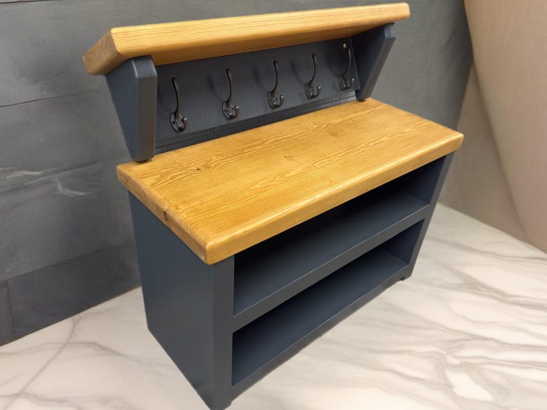Puede incluir: Un organizador de entrada montado en la pared con un estante superior de madera natural y una base gris oscuro. La unidad incluye cinco ganchos de metal negro para colgar objetos y dos compartimentos de almacenamiento abiertos bajo la superficie.