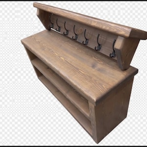 Puede incluir: Un banco de entrada de madera marr&oacute;n con una superficie superior plana, dos estantes de almacenamiento abiertos debajo y un panel trasero con seis ganchos de metal negro para colgar objetos. La unidad est&aacute; hecha de madera con veta visible.