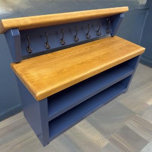 Puede incluir: Un organizador de entrada montado en la pared con un estante superior de madera natural y una superficie de banco de madera a juego. La unidad est&aacute; pintada en azul marino profundo e incluye dos estantes abiertos debajo del banco y seis ganchos de metal.