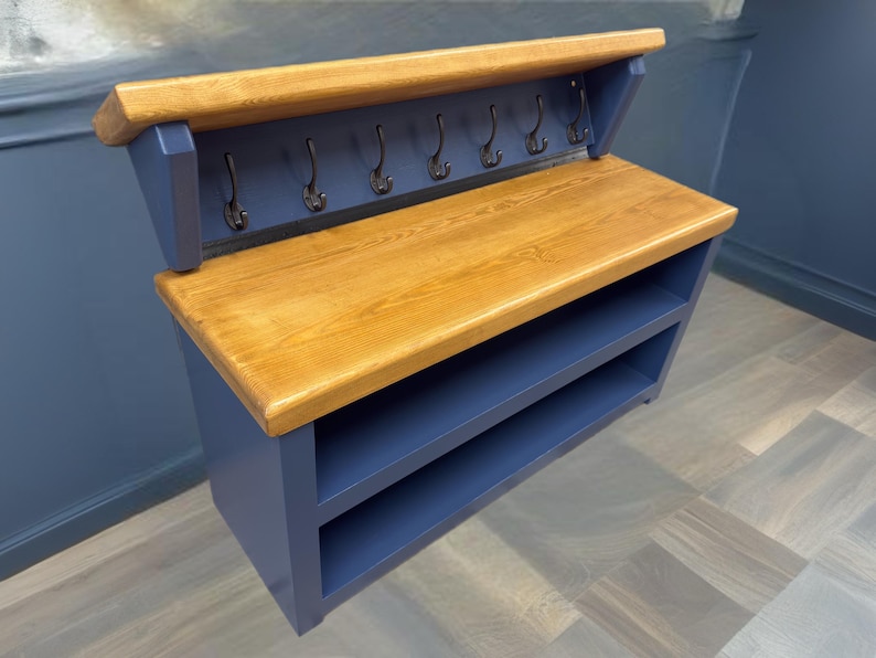 Puede incluir: Un organizador de entrada montado en la pared con un estante superior de madera natural y una superficie de banco de madera a juego. La unidad est&aacute; pintada en azul marino profundo e incluye dos estantes abiertos debajo del banco y seis ganchos de metal.
