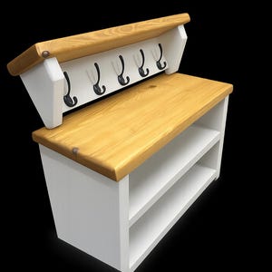 Puede incluir: Un banco de entrada de madera blanca con una superficie superior en tono madera natural, dos estantes de almacenamiento abiertos y un panel trasero con cinco ganchos de metal negro. La unidad tiene una construcci&oacute;n robusta y contrastada.