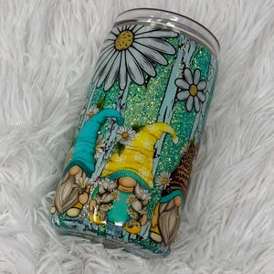 May include: A tumbler with a teal glitter background and a daisy and gnome design. The gnomes wear yellow, teal, and leopard print hats. The tumbler has a clear lid and is decorated with white daisies.