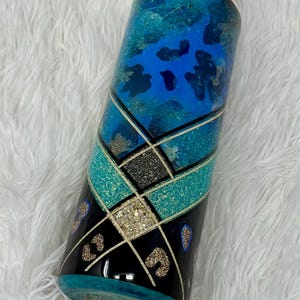 May include: A tall, cylindrical tumbler with a decorative design. The tumbler features a blue and teal color scheme with glitter accents. The design includes geometric shapes and leopard print patterns. The tumbler has a clear lid and a teal base.