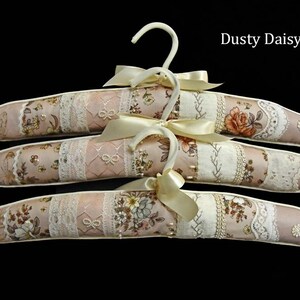 May include: Three padded clothes hangers with floral and lace fabric in shades of pink, cream, and beige. Each hanger has a cream-coloured bow and hook. The text "Dusty Daisy" is in the upper right corner.
