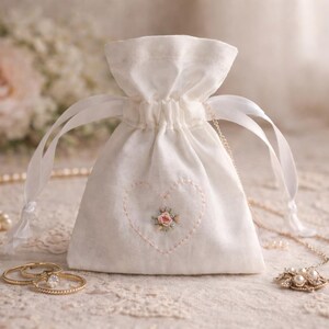 May include: A cream-coloured drawstring bag with a heart-shaped floral embroidery. The bag is tied with white ribbons and sits on a lace surface, next to gold rings and a pearl necklace. A rose and baby's breath are in the background.