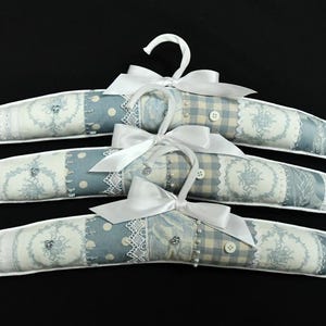 May include: Three padded clothes hangers with a light blue and white floral and polka dot pattern. Each hanger has a white satin bow and a white hook. The hangers are decorated with lace and small buttons.