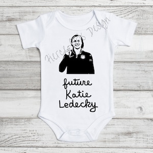 Pode incluir: Macacão branco para bebê com uma imagem em preto e branco de uma nadadora segurando uma medalha de ouro. O texto "future Katie Ledecky" está impresso abaixo da imagem.