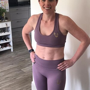 May include: A mauve-colored sports bra and leggings set. The bra and leggings have a ribbed texture and a gold logo. The woman is wearing a gray headband and a black smartwatch. The outfit is designed for fitness activities.
