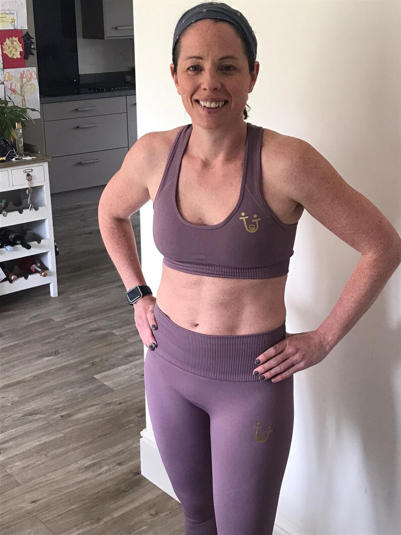 May include: A mauve-colored sports bra and leggings set. The bra and leggings have a ribbed texture and a gold logo. The woman is wearing a gray headband and a black smartwatch. The outfit is designed for fitness activities.
