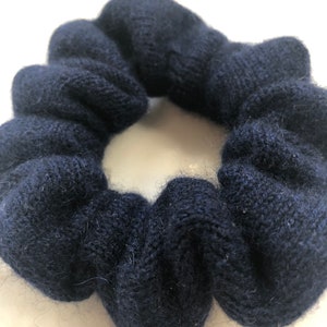 May include: A navy blue scrunchie made of a soft, knitted material. The scrunchie is circular and has a gathered appearance. The dark blue color is consistent throughout the fabric.
