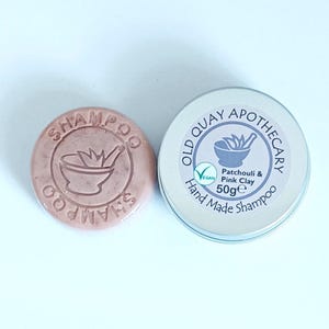 May include: A pink shampoo bar with the word "SHAMPOO" embossed around a mortar and pestle design. Beside it is a silver tin with the text "OLD QUAY APOTHECARY" and "Patchouli & Pink Clay Hand Made Shampoo" with a vegan symbol. The tin contains 50g of product.