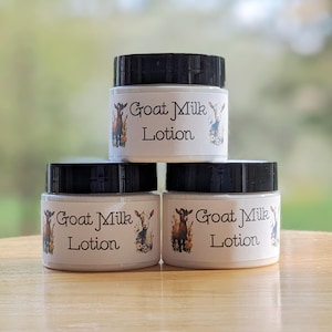 May include: Three small jars of goat milk lotion are stacked. Each jar has a black lid and a white label with the words "Goat Milk Lotion" and a goat illustration. The jars are on a wooden surface.