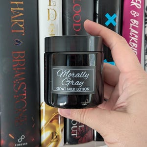 May include: A dark gray jar of goat milk lotion with a black lid. The label reads "Morally Gray GOAT MILK LOTION" in elegant script. The jar is held in front of a bookshelf with book titles visible.