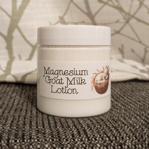 May include: A white jar of Magnesium Goat Milk Lotion with a white lid. The label features the text "Magnesium Goat Milk Lotion" and a brown illustration of a sleeping baby goat. The jar is sitting on a gray surface.