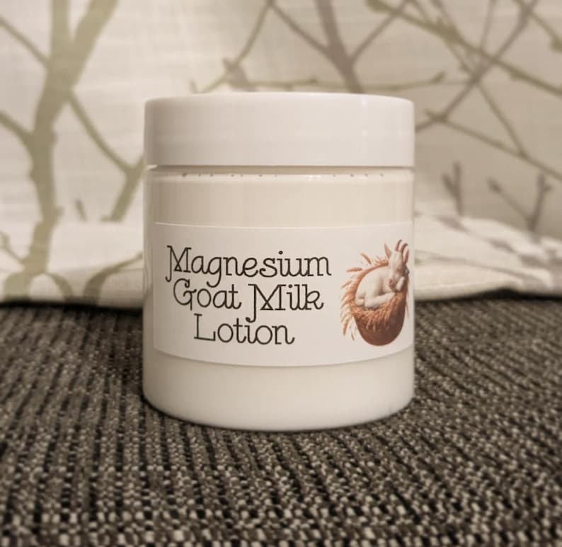 May include: A white jar of Magnesium Goat Milk Lotion with a white lid. The label features the text "Magnesium Goat Milk Lotion" and a brown illustration of a sleeping baby goat. The jar is sitting on a gray surface.