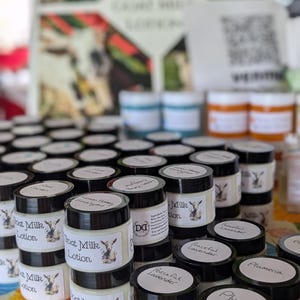 May include: A display of small jars of goat milk lotion with black lids and white labels. The labels feature a goat illustration and the words "Goat Milk Lotion." Various scents are labeled, including "Peaceful Lavender" and "Plumeria."
